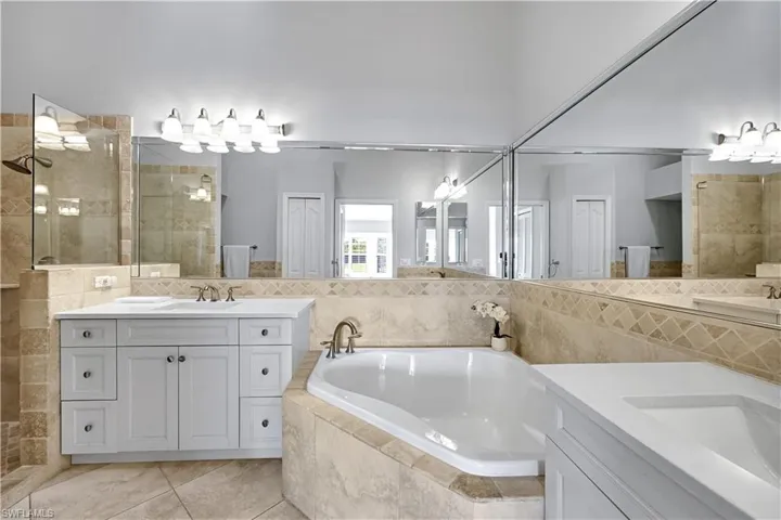 Bathroom featuring white cabinetry, dual sinks with light-toned solid surface countertops, a corner soaking tub with tile surround, expansive walk-in shower, and an expansive wall-to-wall vanity mirror