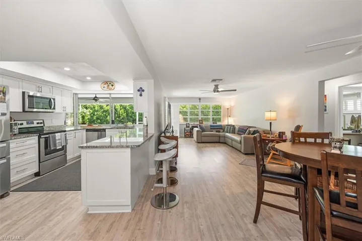 Open-concept living area featuring wood-finish flooring throughout, a kitchen with white cabinetry, granite-style countertops, stainless steel appliances, and a breakfast bar