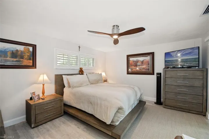 Bedroom featuring a floating ceiling fan with integrated lighting, wood-finish flooring, and a window with white plantation shutters