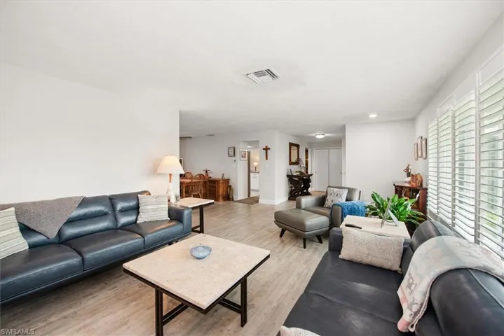 Spacious living area featuring light wood-finish flooring, white walls, and a large window with plantation shutters