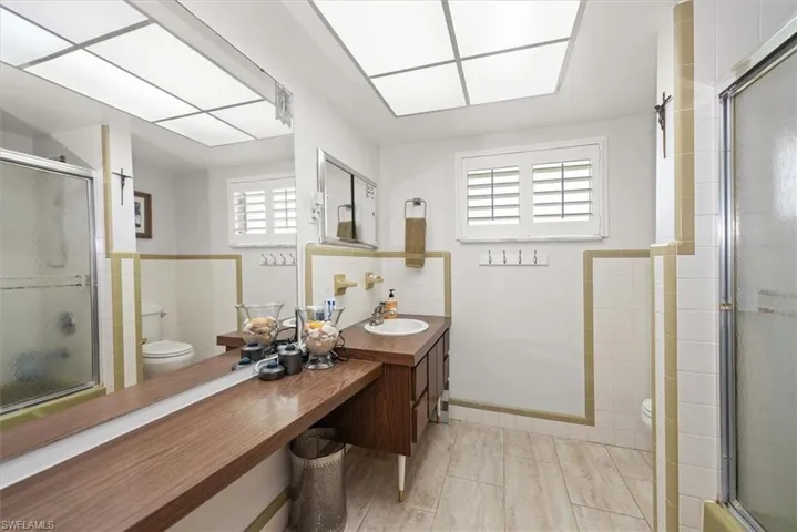 Bathroom featuring wood-finish flooring, a vanity with wood-tone cabinetry, a recessed ceiling light fixture, white tile walls with a gold-toned trim, and a window with white louvered shutters
