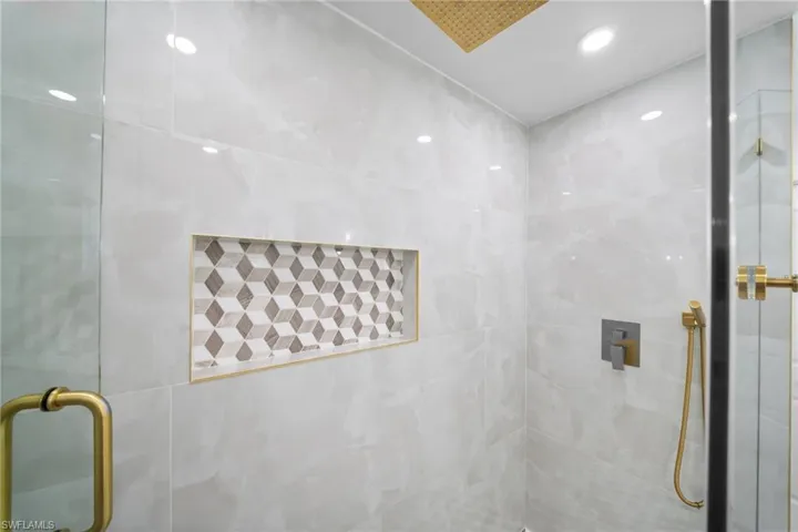 Walk-in shower featuring large-format stone-finish tile, geometric-patterned tile niche, brass-finish hardware, handheld shower fixture, and a glass door enclosure