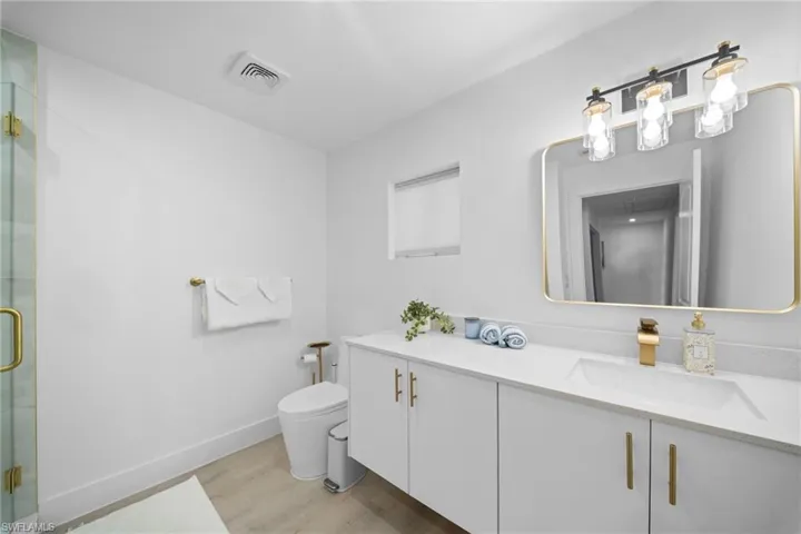 3rd full bathroom. Modern vanity with white cabinetry, rectangular basin, and metallic gold hardware, complemented by a gold-framed wall mirror, multi-bulb vanity light, and glass-enclosed shower