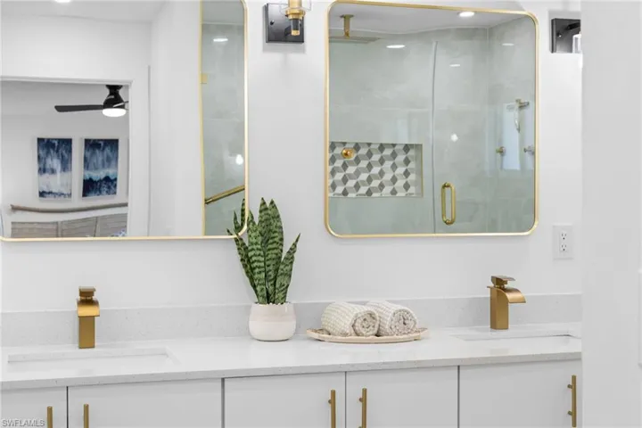 Double vanity featuring a light-toned countertop, rectangular undermount sinks, gold-finish hardware, two gold-framed mirrors, and a glass-enclosed shower with a geometric tile inlay