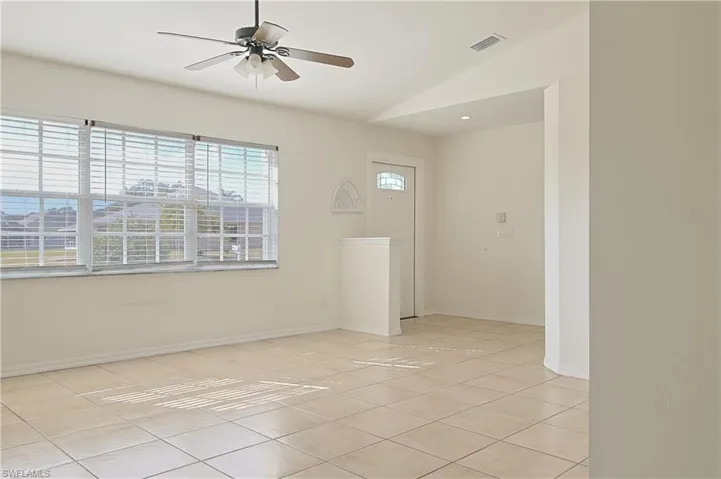 Spacious interior room featuring light-toned square tile flooring, multiple windows with horizontal blinds, a ceiling fan with integrated lighting, recessed ceiling lighting, and a white entry door with a decorative glass insert