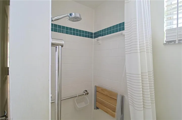 Shower with white subway tile, an accent band of teal square tiles, a handheld shower head, a chrome grab bar, and a folding wood-slat shower seat