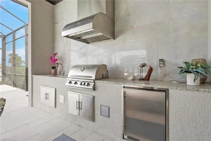 Outdoor kitchen featuring a built-in stainless steel grill, integrated range hood, under-counter refrigerator, and a sink with a stainless steel faucet