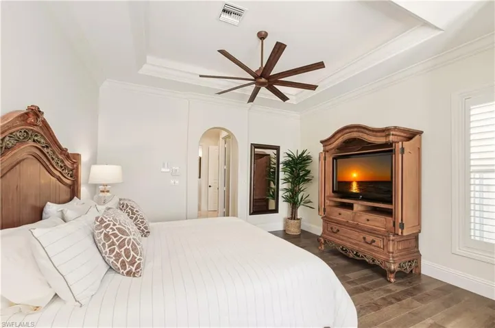 Bedroom featuring a raised ceiling, dark wood-type flooring, ceiling fan, arched walkways, and crown molding
