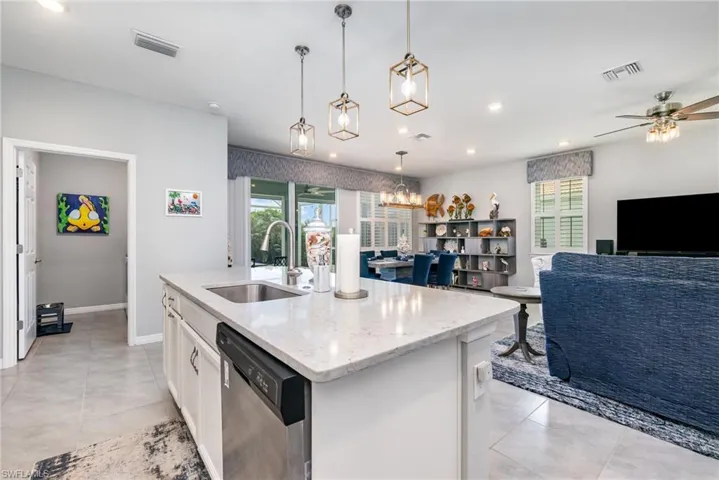 Kitchen featuring stainless steel dishwasher, white cabinets, light stone countertops, a center island with sink, and hanging light fixtures