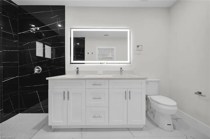 Bathroom featuring a dual-sink vanity with white cabinetry, a rectangular illuminated mirror, and a walk-in shower with black tile and chrome fixtures