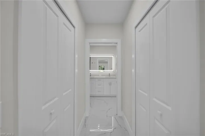 Hallway featuring white bi-fold doors, light neutral wall paint, and marble-look tile flooring leading to a bathroom with a white vanity and illuminated mirror