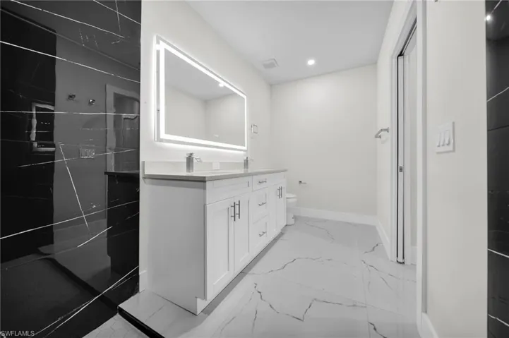 Black-tiled shower enclosure with linear marbling, dual sink vanity with stone countertop, backlit rectangular mirror, recessed lighting, and large format floor tiles with linear marbling
