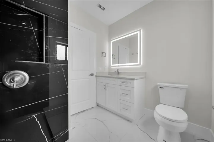 Bathroom featuring a black marble-style shower with a built-in niche, white vanity with a light countertop, illuminated mirror, and white tile flooring