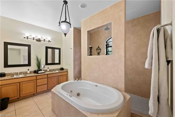 Bathroom featuring an integrated jetted tub, a separate tiled shower with a rain shower head and recessed niche, and a dual vanity with light wood cabinetry and stone countertops