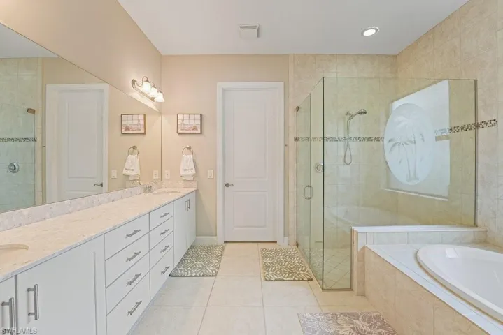 Dual vanity with light-toned countertops, tiled flooring, glass-enclosed shower with decorative inlay, separate soaking tub, and full-length wall mirror