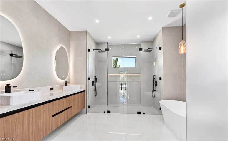 This bathroom features a double vanity with two vessel sinks, back-lit mirrors, and a dual-head shower with a glass enclosure