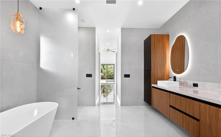 Contemporary bathroom featuring a freestanding white bathtub, a large vanity with a white countertop and wood cabinetry, and a unique backlit mirror