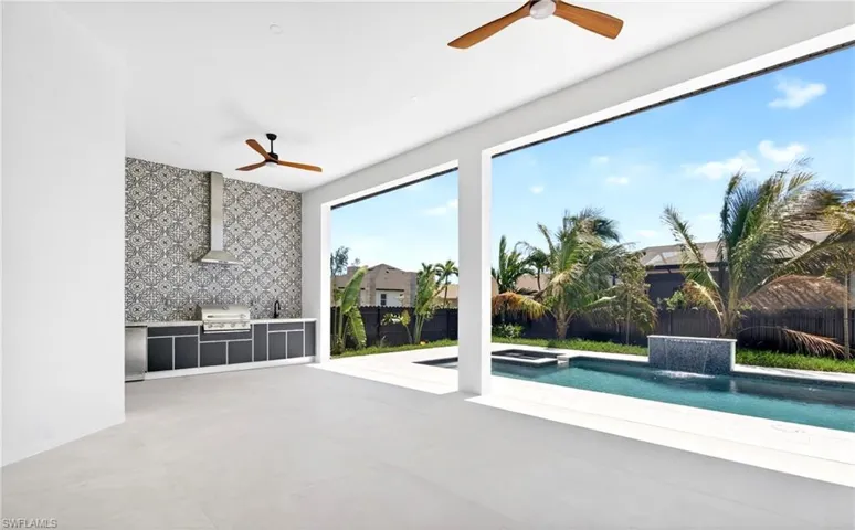 Expansive covered patio featuring an outdoor kitchen with a patterned tile backsplash, stainless steel grill, and an overhead extractor fan, with a view of the pool and palm trees