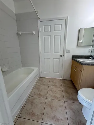 Full bathroom featuring a white bathtub with tile surround, a light wood-finish vanity with a dark countertop, a white toilet, and square floor tiles