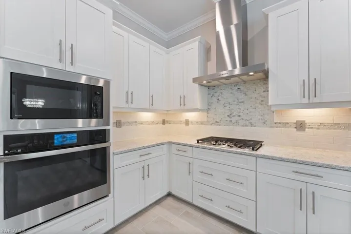 Kitchen featuring wall chimney range hood, stainless steel appliances, crown molding, white cabinets, and light stone countertops