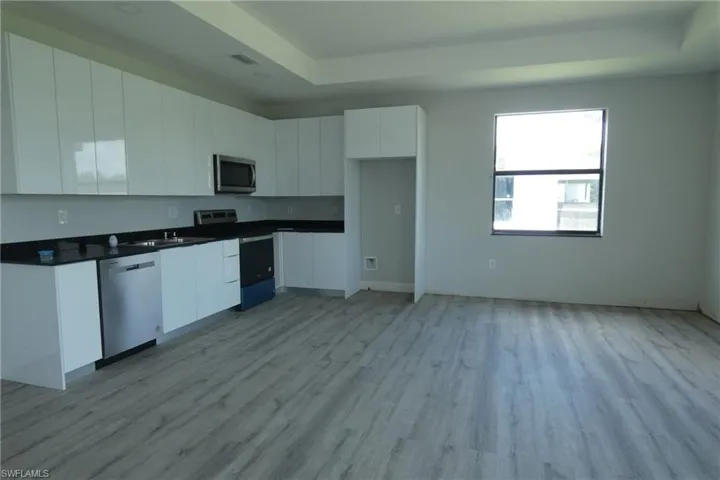 Open-concept kitchen featuring white gloss cabinetry, dark stone countertops, stainless steel appliances, wood-finish flooring, and a recessed ceiling detail