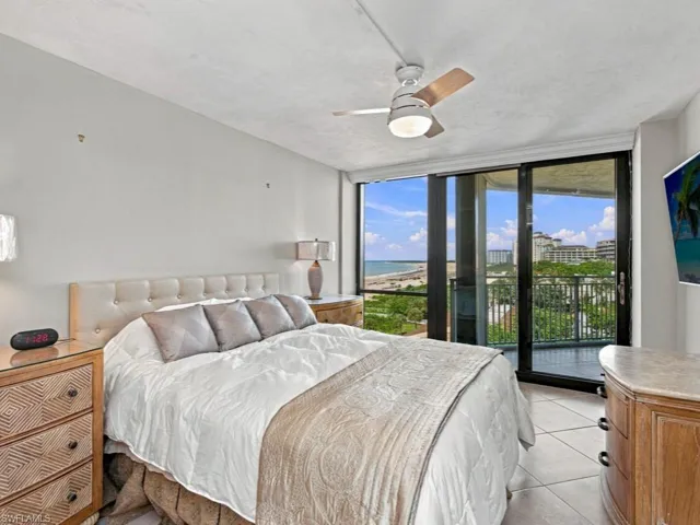 Floor-to-ceiling sliding glass doors, white tile flooring, ceiling fan with integrated light, and access to a private balcony overlooking the coastline