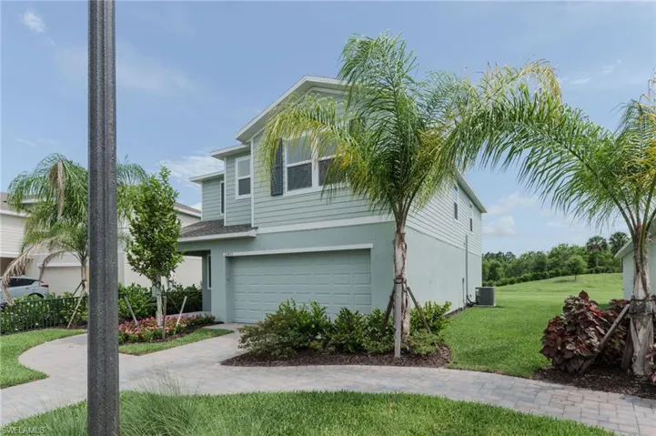 Contemporary two-story exterior featuring a light green and gray facade, an integrated garage, a paver driveway, mature palm trees, and lush landscaping