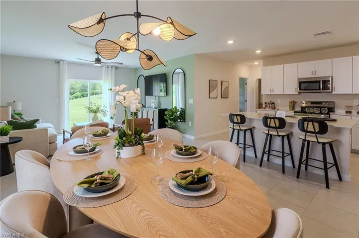 Open-concept living space featuring a dining area with a distinctive chandelier, a contemporary kitchen with white cabinetry, a kitchen island with seating, stainless steel appliances, and a living area with sliding glass doors opening to a green exterior
