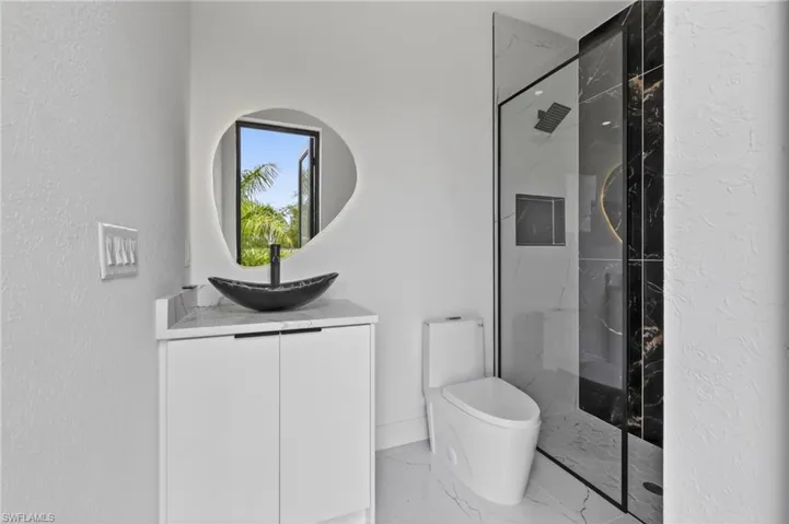 Modern bathroom featuring a glass-enclosed shower with black-veined wall tile, an irregular-shaped illuminated mirror, and a black vessel sink set on a white vanity with a light-colored countertop