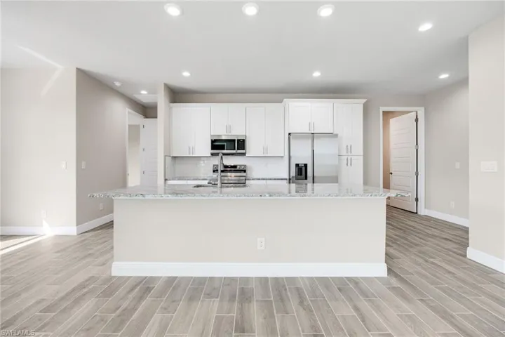 Kitchen featuring light stone countertops, appliances with stainless steel finishes, white cabinets, a kitchen island with sink, and recessed lighting-Finishes may vary