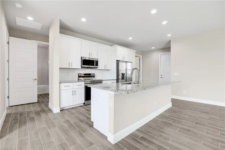Kitchen with stainless steel appliances, white cabinetry, wood tiled floors, an island with sink, and recessed lighting-Finishes may vary
