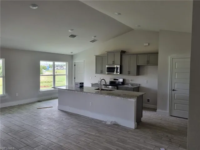Kitchen featuring light wood-type flooring, gray cabinets, vaulted ceiling, appliances with stainless steel finishes, and a center island with sink