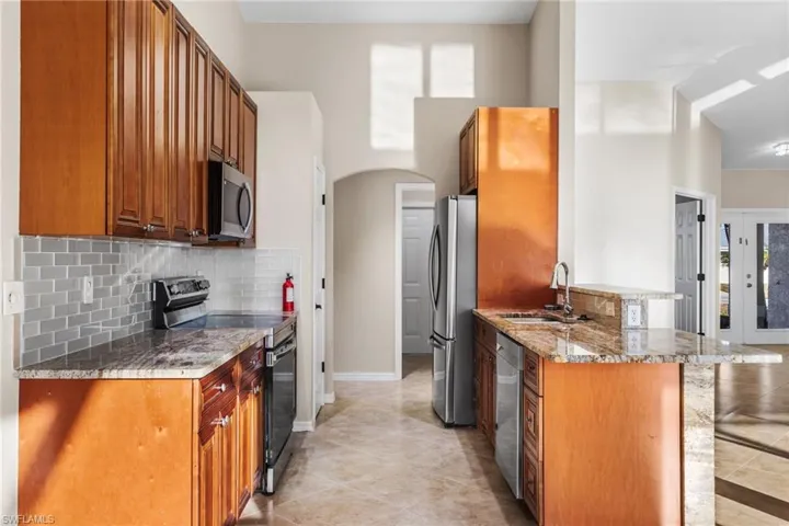 Kitchen featuring stainless steel appliances, arched walkways, light stone counters, wood finish cabinetry, and a high ceiling