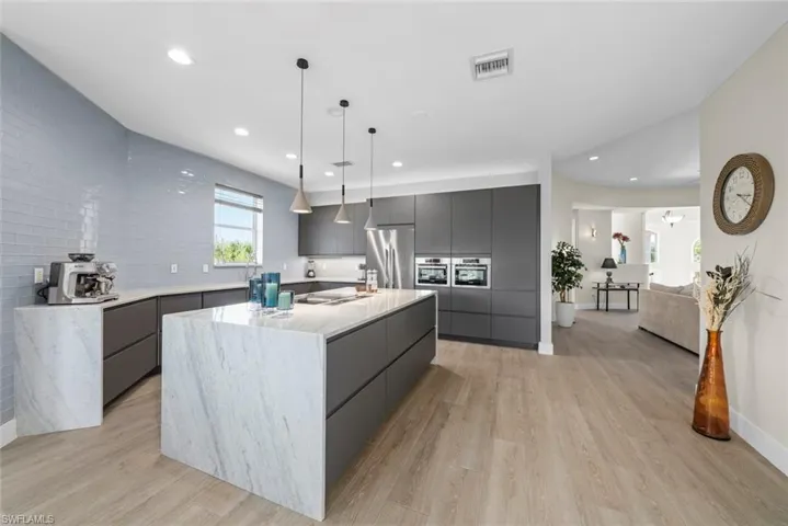 Contemporary kitchen featuring a large waterfall island with a stone surface, sleek dark cabinetry, integrated stainless steel appliances, a light blue subway tile backsplash, and wood-finish flooring