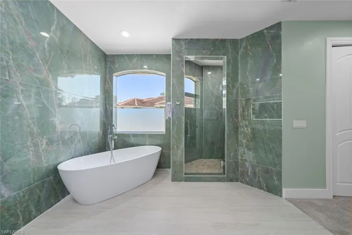 Bathroom featuring a freestanding soaking tub, a walk-in shower with glass enclosure, large format green marble-look wall tiles, light-toned floor tiling, and an arched window