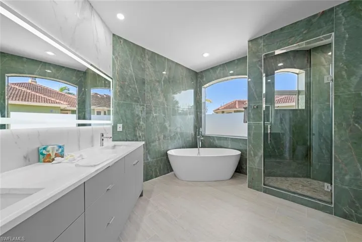 Spacious bathroom featuring green marble-look wall tiling, a freestanding soaking tub, a glass-enclosed shower, a dual vanity with a light gray cabinet, and light-toned tile flooring