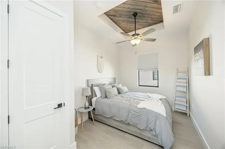 Recessed ceiling with wood-finish paneling, ceiling fan with integrated lighting, wood-finish flooring, white flat-panel door with black hardware, and window with roller shade