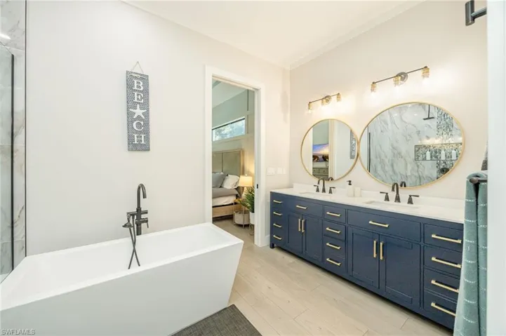 Spacious bathroom featuring a freestanding soaking tub with a floor-mounted faucet, a dual vanity with a dark blue cabinet base, gold hardware, and round mirrors, and wood-finish flooring
