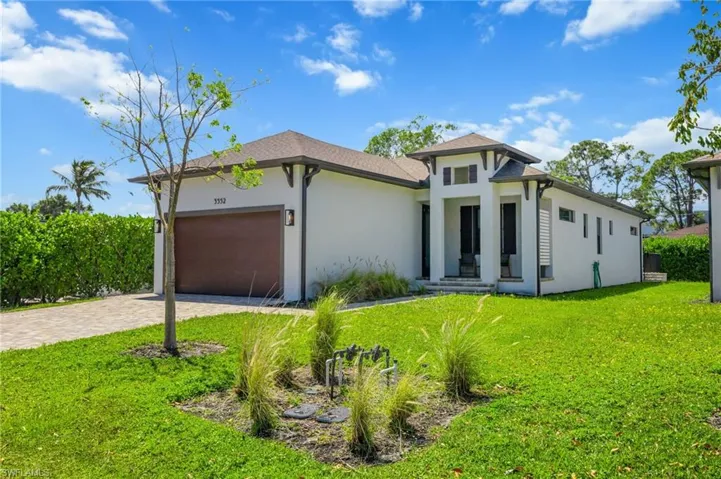Contemporary stucco exterior with a prominent entry tower, dark trim accents, a paver driveway, an integrated garage with a wood-finish door, and manicured green landscaping