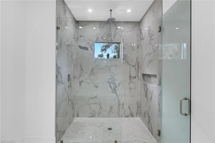 Spacious walk-in shower featuring floor-to-ceiling marbled tile, clear glass enclosure, built-in recessed shelf, and a ceiling-mounted rain shower head