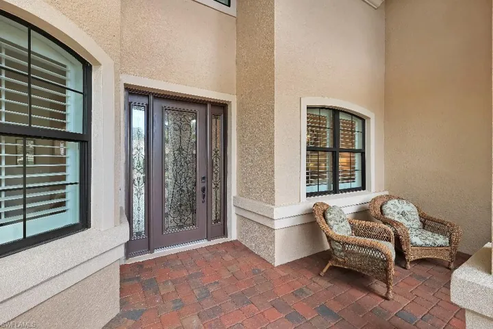 Covered entry featuring a decorative glass main door with sidelights, arched windows with plantation shutters, brick paver flooring, and stucco exterior finishes
