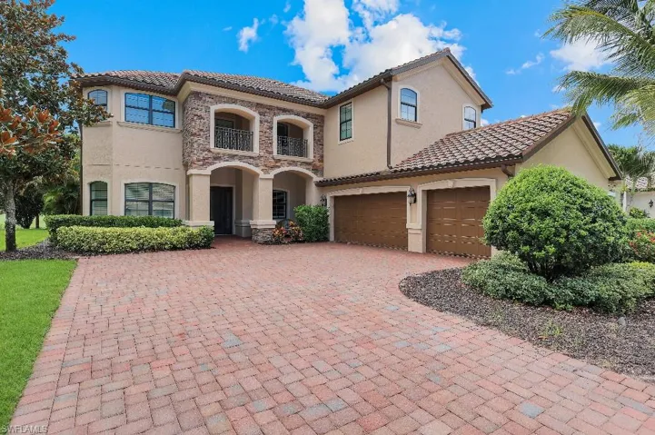 Mediterranean-inspired stucco exterior featuring a terra cotta tile roof, stone facade accents, decorative wrought iron balcony railings, and a red brick paver driveway
