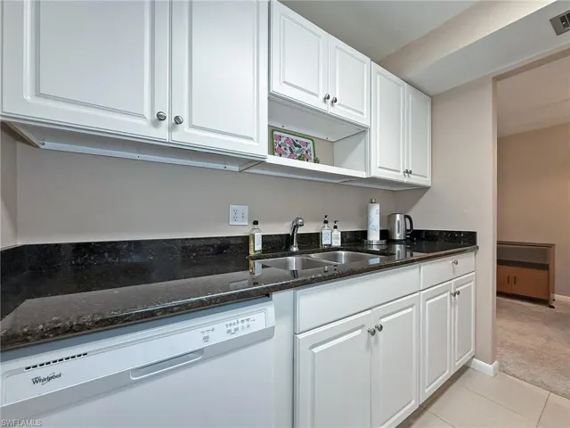 Kitchen with a sink, white dishwasher, dark stone countertops, light colored carpet, and white cabinets