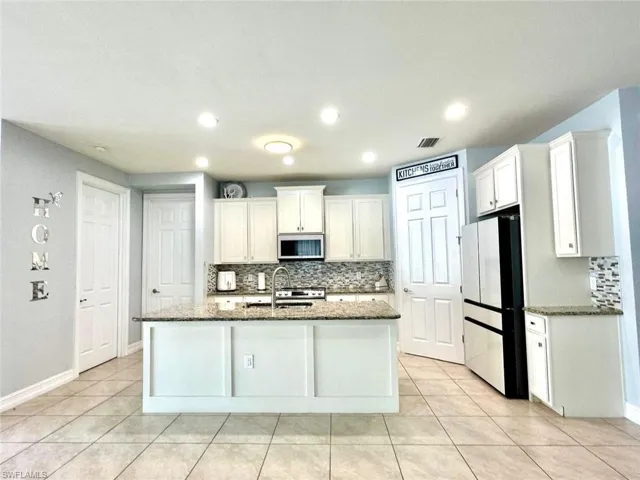 Kitchen with light tile patterned floors, tasteful backsplash, dark stone counters, freestanding refrigerator, and white cabinetry
