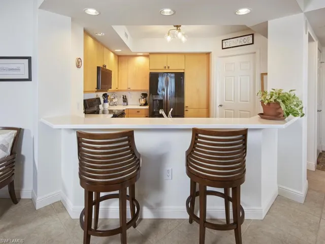 Kitchen with light tile patterned floors, kitchen peninsula, black appliances, light brown cabinetry, and a kitchen bar