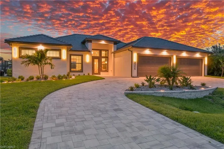 Contemporary stucco exterior featuring a two-car garage with overhead lighting, a paver driveway, manicured landscaping, and a tiled roof