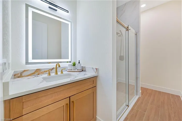 Bathroom vanity featuring a backlit mirror, gold-finish fixtures, light wood-finish cabinetry, and a white countertop with amber veining