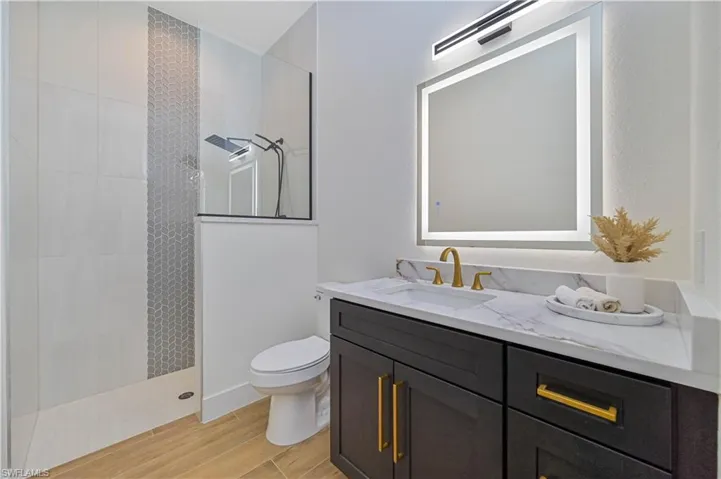 Bathroom featuring a walk-in shower with glass enclosure, decorative tile accent, wood-finish flooring, a vanity with dark cabinetry, and an illuminated mirror