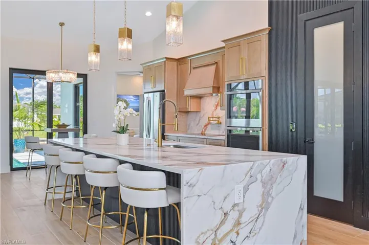 Spacious kitchen featuring a large island with waterfall countertop, stainless steel appliances, light wood cabinetry, and a black paneled accent wall