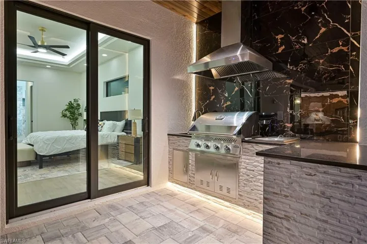 Outdoor kitchen featuring a built-in stainless steel grill, stone veneer base, black stone countertops, and a black and gold marble-patterned backsplash with integrated lighting
